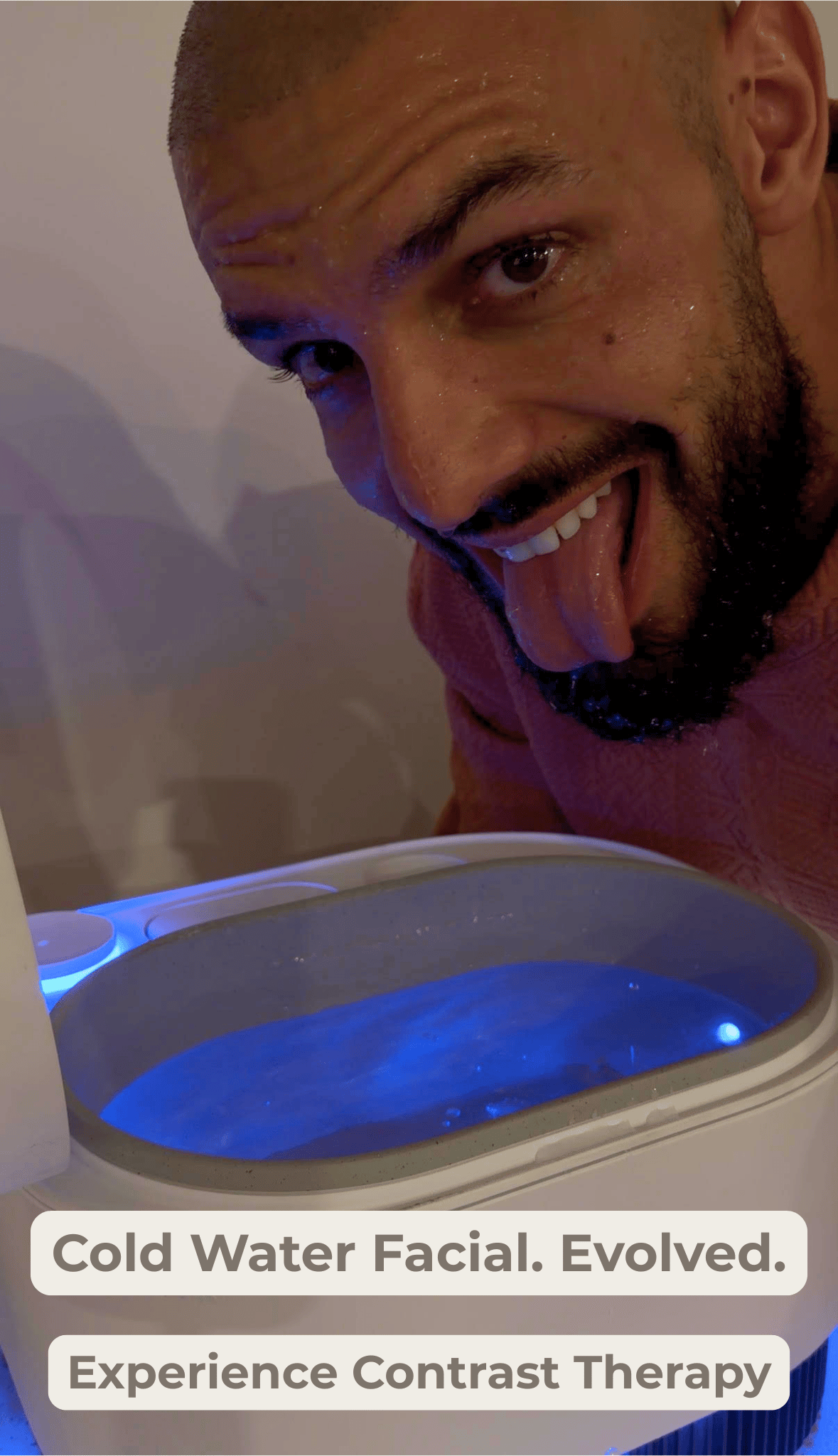 A close-up lifestyle shot of a man with a shaved head and beard sticking out his tongue in a playful manner next to a Frosteam device filled with ice  water